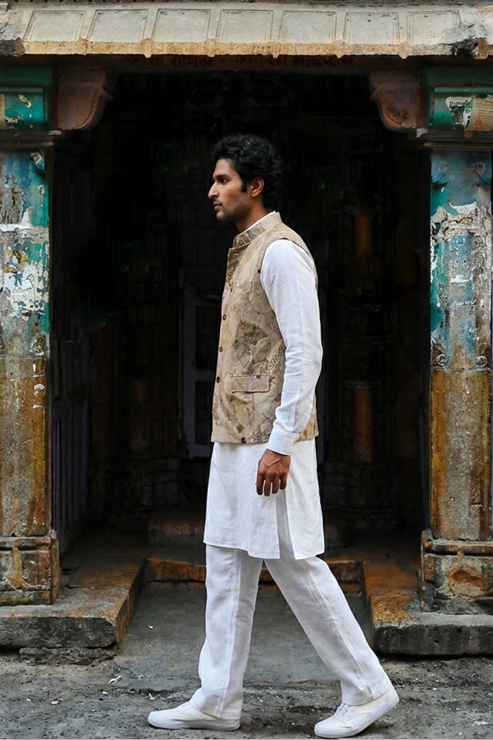 A man in a traditional Indian outfit consisting of a white kurta and trousers with a beige eco-printed hemp vest, walking past a rustic, weathered doorway.