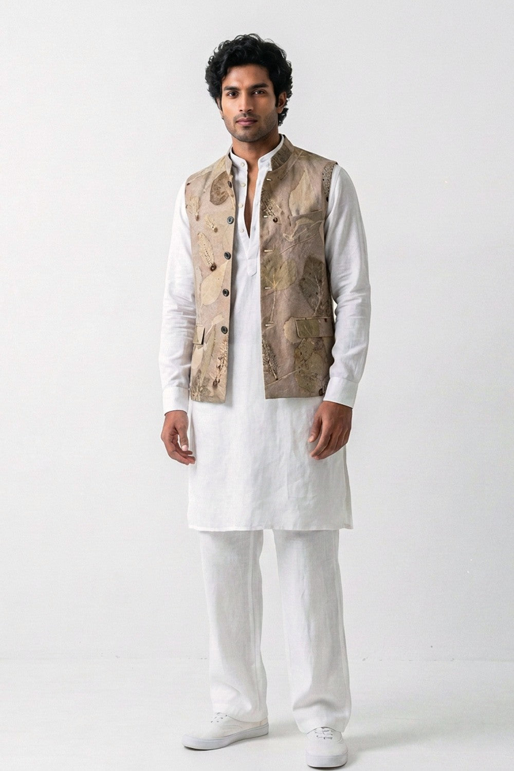 A man modeling a white traditional outfit consisting of a kurta, trousers, and white sneakers, paired with a beige hemp eco-printed vest over a white shirt, against a plain white studio background.