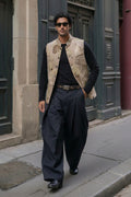 A man walking on a city street wearing a beige eco-printed patterned vest over a black long-sleeve shirt, black wide-leg trousers, and black shoes.