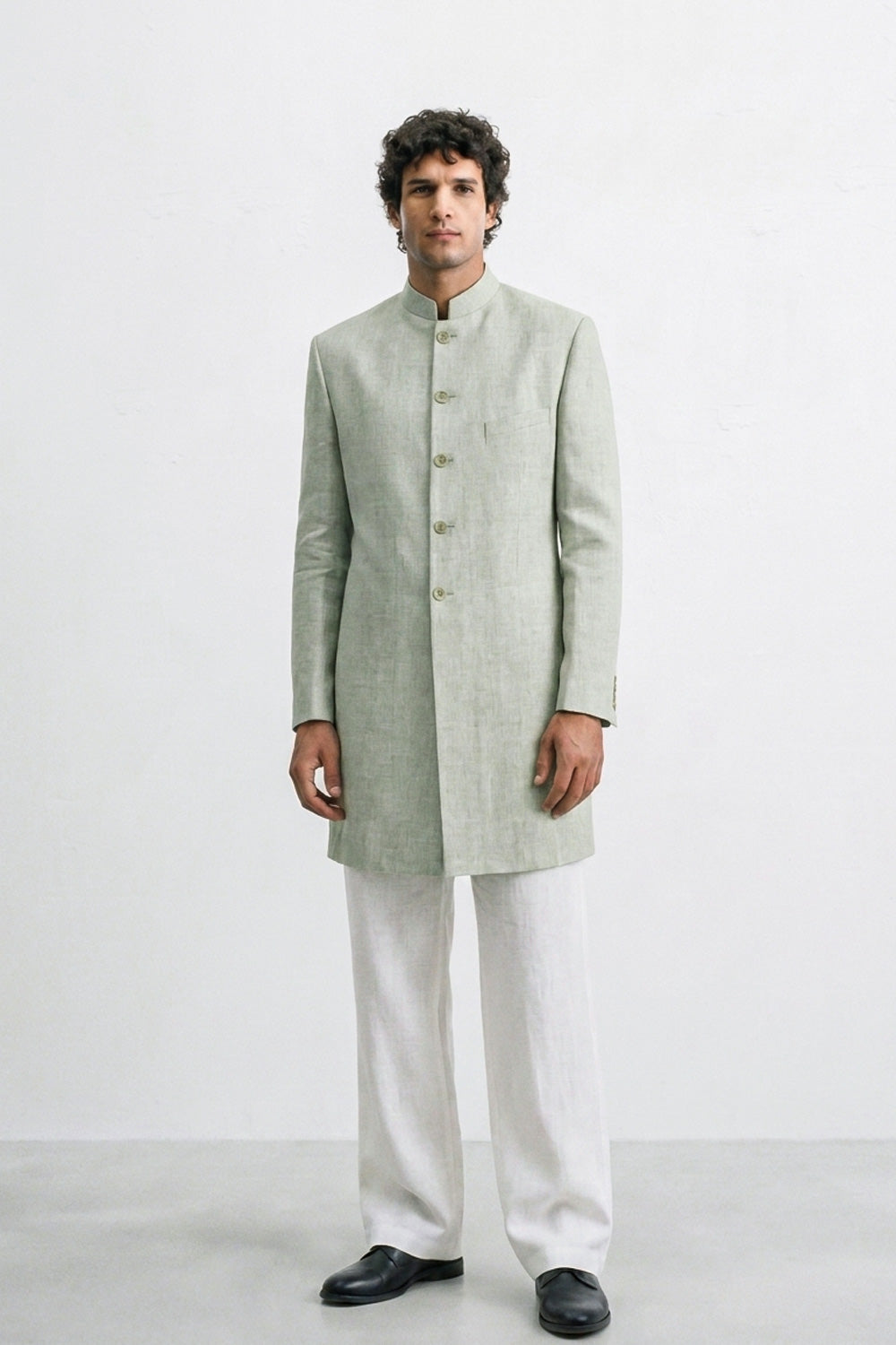 A man modeling a light sage green linen sherwani jacket with white trousers and black shoes, standing against a plain white studio background.