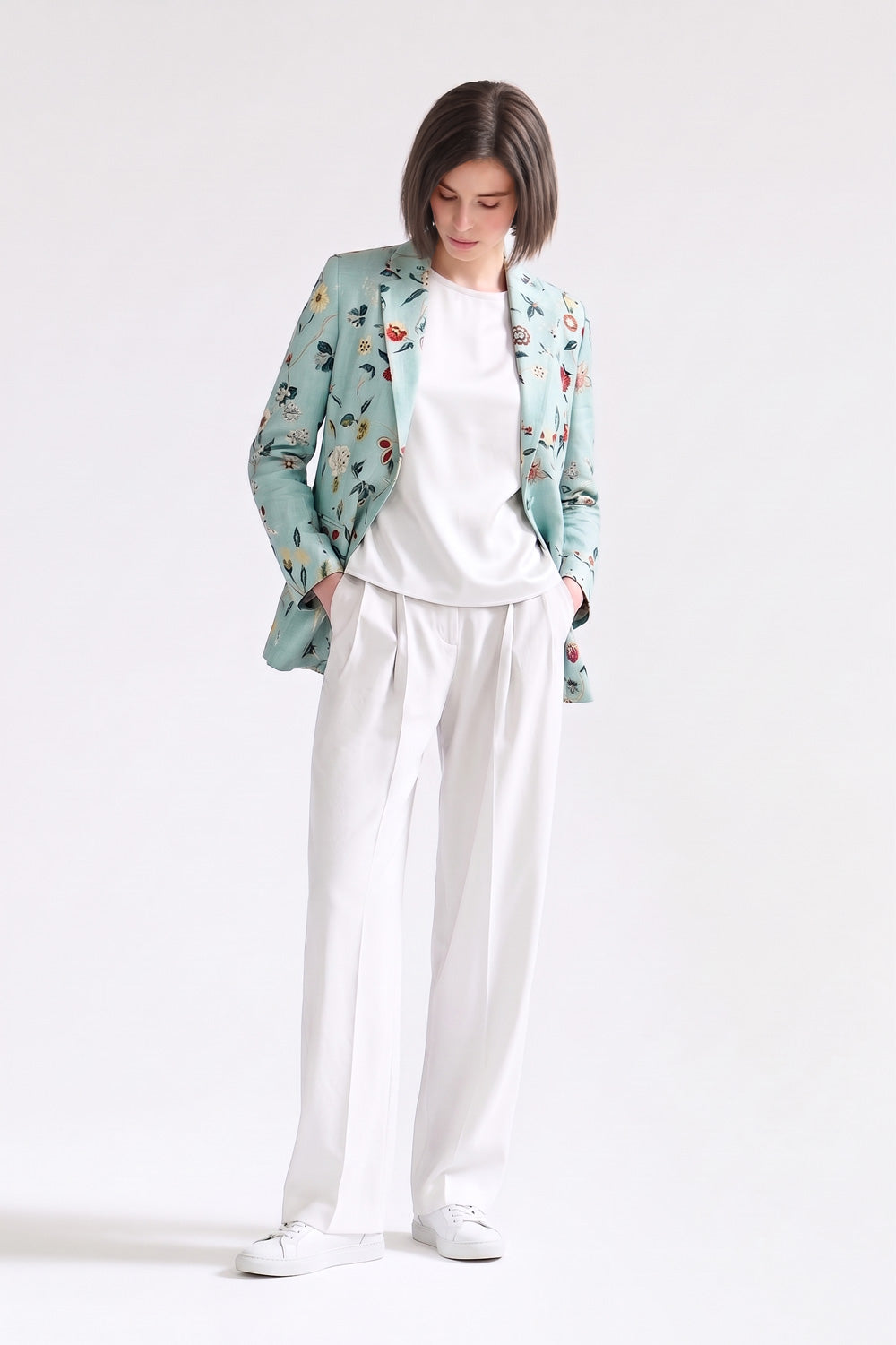A model stands against a plain white studio background wearing a mint green floral patterned linen blazer over a white t-shirt and white wide-leg trousers.
