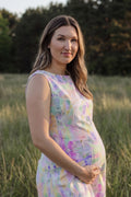 A pregnant woman wearing a sleeveless pastel tie-dye maternity hemp dress stands in a grassy field at sunset.