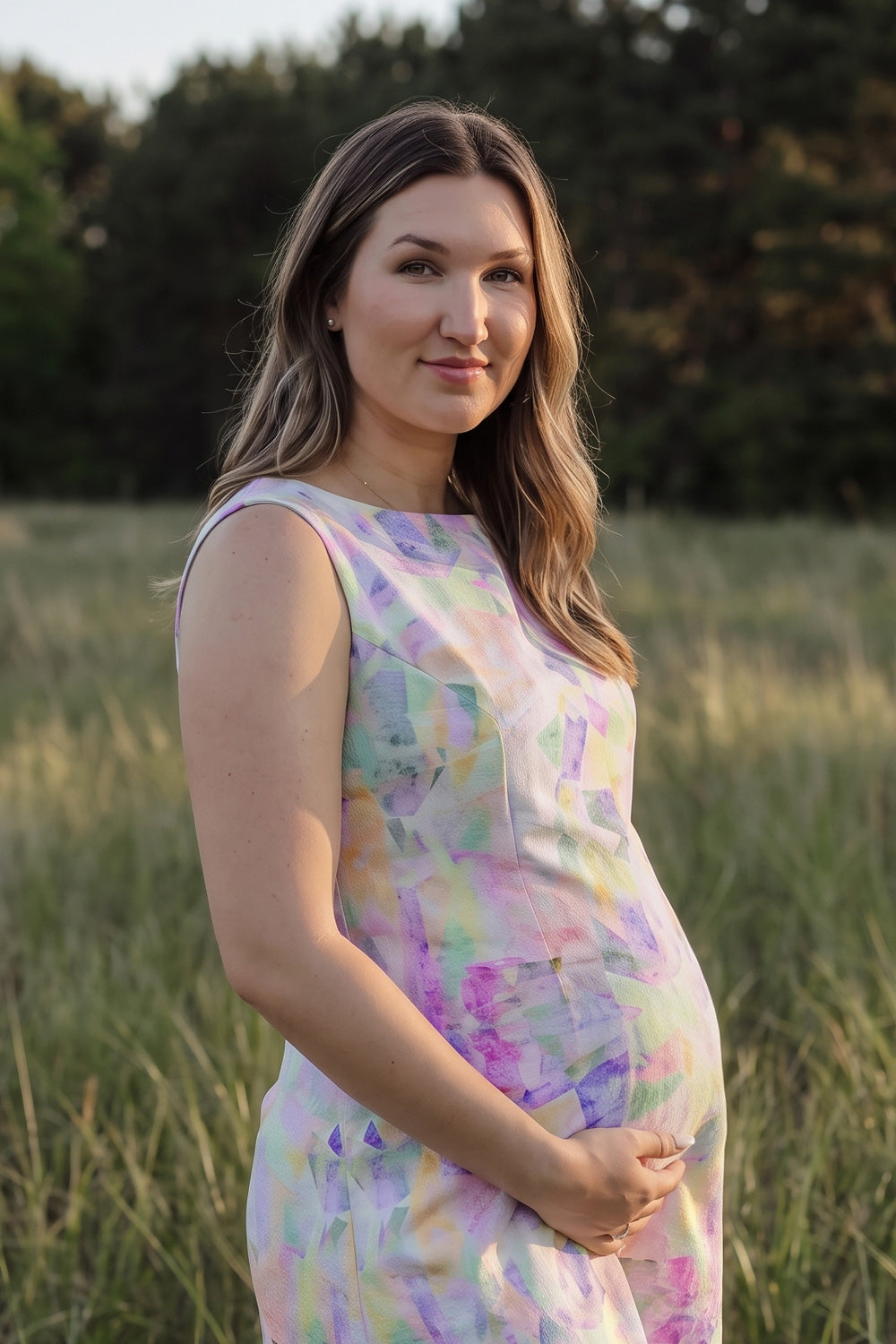 A pregnant woman wearing a sleeveless pastel tie-dye maternity hemp dress stands in a grassy field at sunset.