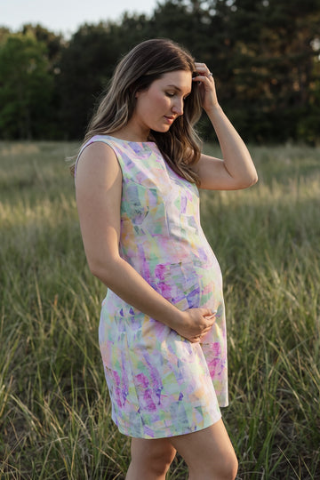 A pregnant woman wearing a sleeveless, knee-length dress with a pastel multicolor watercolor pattern, standing in a grassy field at sunset.