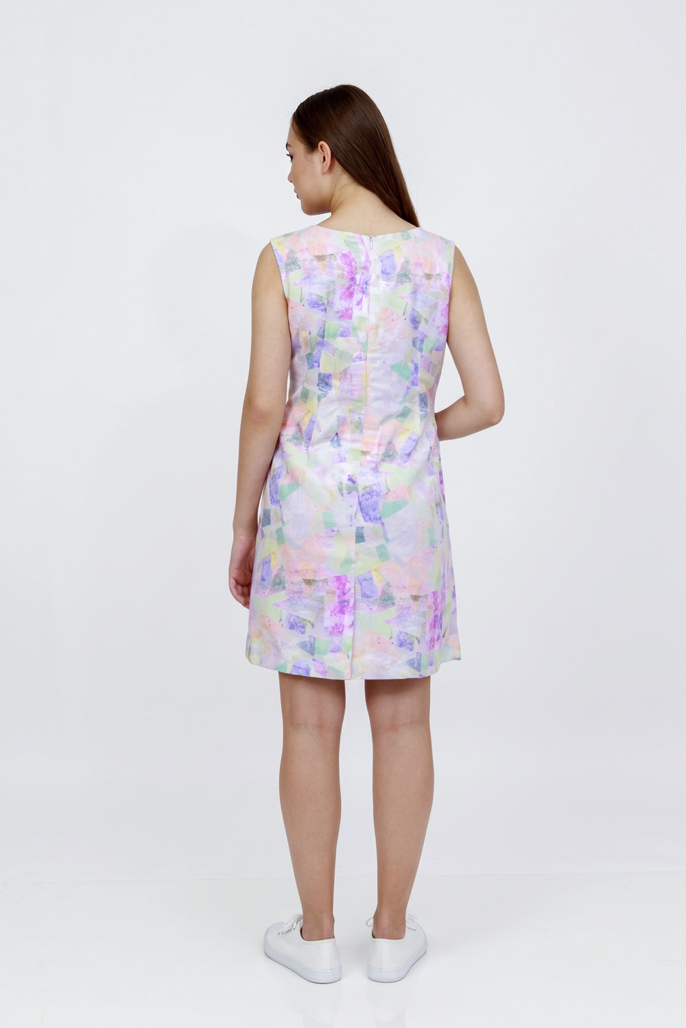 A woman viewed from the back wearing a sleeveless A-line hemp dress with a pastel multicolor floral print, paired with white sneakers, against a plain white studio background.