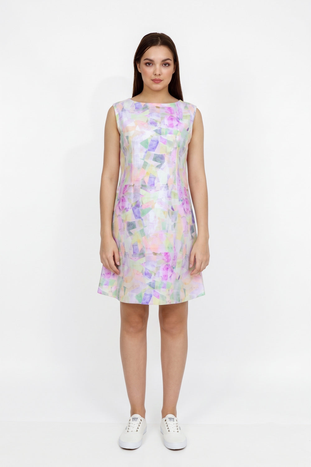 A woman modeling a sleeveless A-line  dress with an abstract pastel watercolor pattern in purple, pink, green, and blue on a white base, paired with white sneakers, against a plain white studio background.