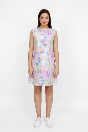 A woman modeling a sleeveless A-line  dress with an abstract pastel watercolor pattern in purple, pink, green, and blue on a white base, paired with white sneakers, against a plain white studio background.