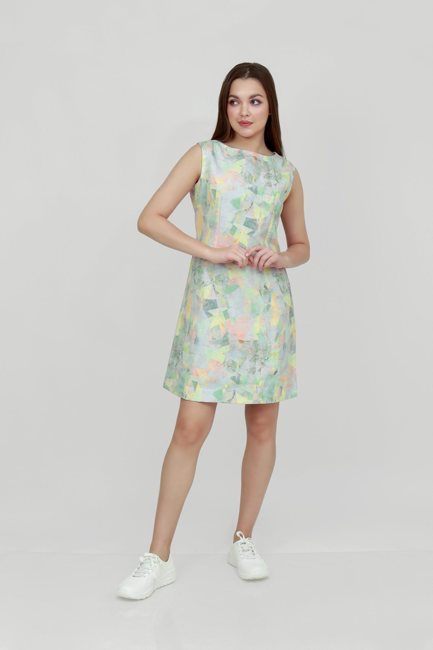 A woman standing in a studio setting, wearing a mid-length floral hemp seersucker dress with a boat neckline and sleeveless top, paired with white sneakers.
