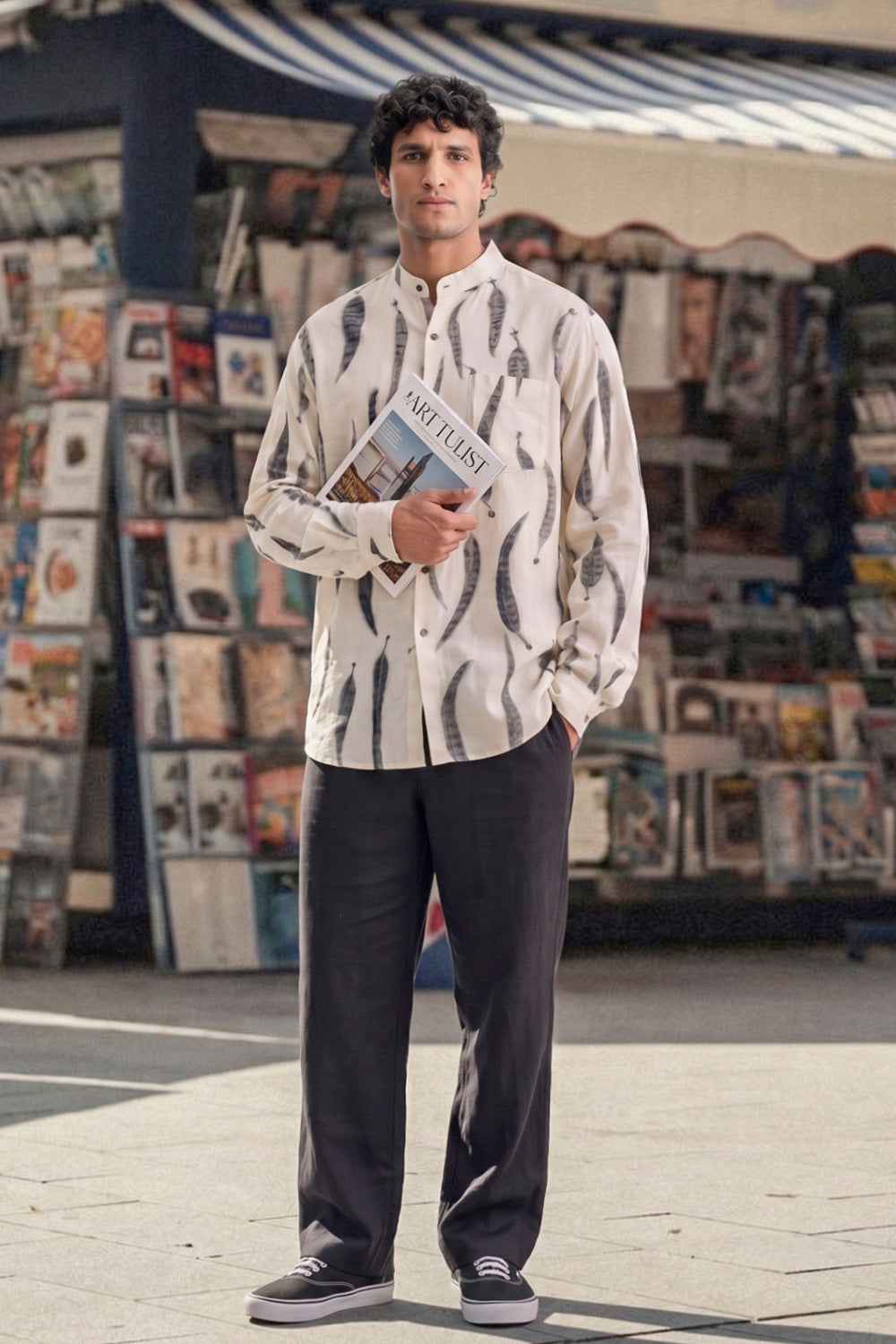 A man stands outdoors in front of a market stall, wearing a cream-colored long-sleeve button-up shirt with a black leaves eco-print pattern, dark gray trousers, and black sneakers.