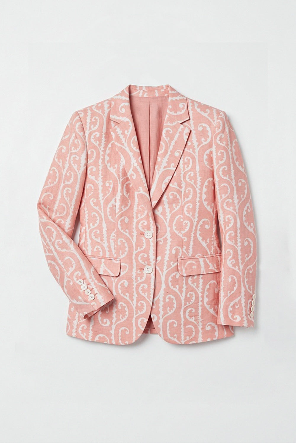A woman stands against a plain white studio background wearing a peach-pink linen blazer with a white ornamental pattern, white wide-leg pants, and white heeled sandals.