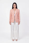 A woman stands against a plain white studio background wearing a peach-pink linen blazer with a white ornamental pattern, white wide-leg pants, and white heeled sandals.
