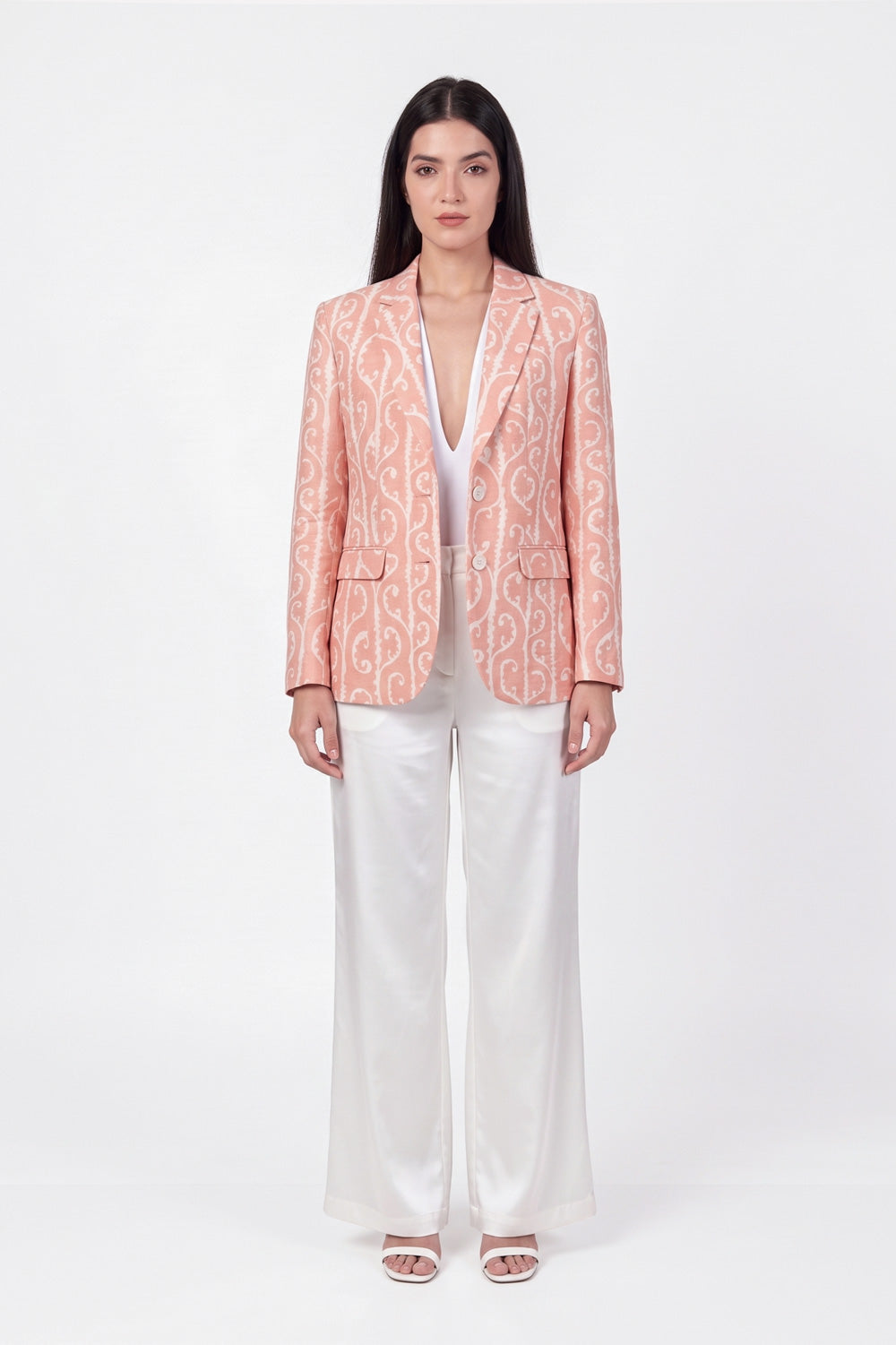 A woman stands against a plain white studio background wearing a peach-pink linen blazer with a white ornamental pattern, white wide-leg pants, and white heeled sandals.