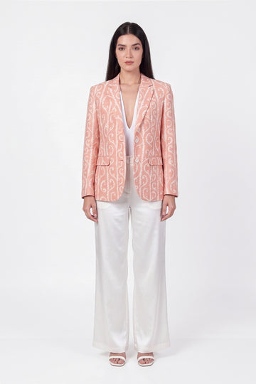 A woman stands against a plain white studio background wearing a peach-pink linen blazer with a white ornamental pattern, white wide-leg pants, and white heeled sandals.