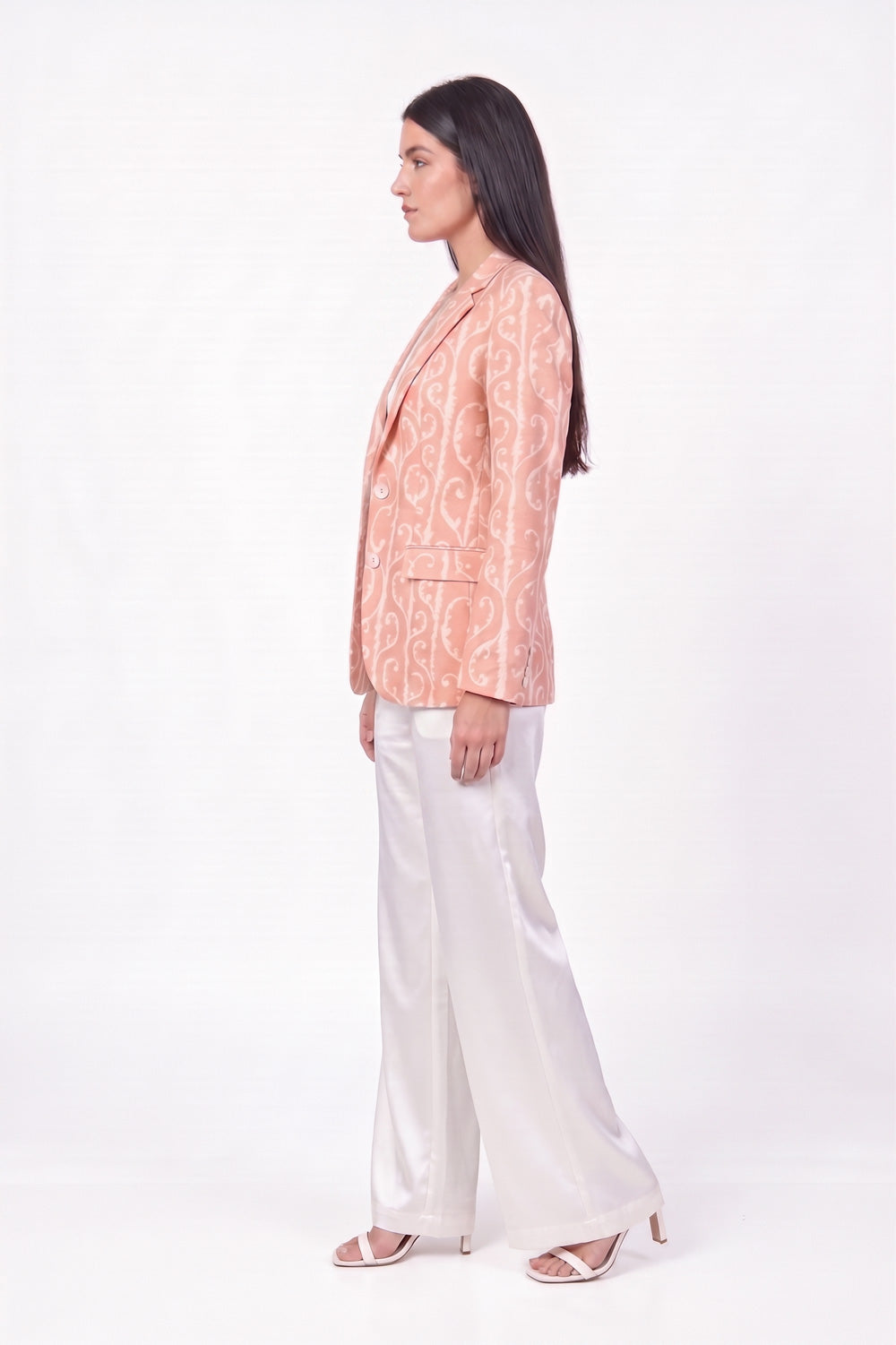 A woman stands in profile wearing a peach-colored printed linenblazer with a white paisley pattern, paired with white satin wide-leg trousers and clear high-heeled sandals, set against a plain white studio background.