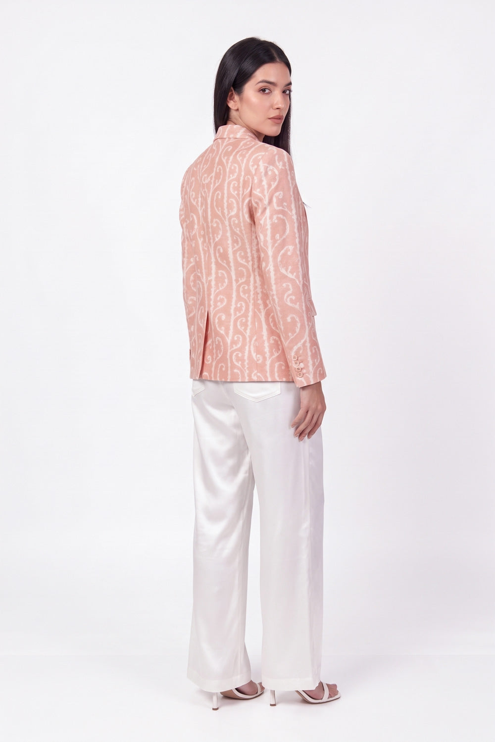 A woman shown from the back wearing a peach and white patterned long-sleeve linen blazer with white satin wide-leg trousers, standing against a plain white studio background.