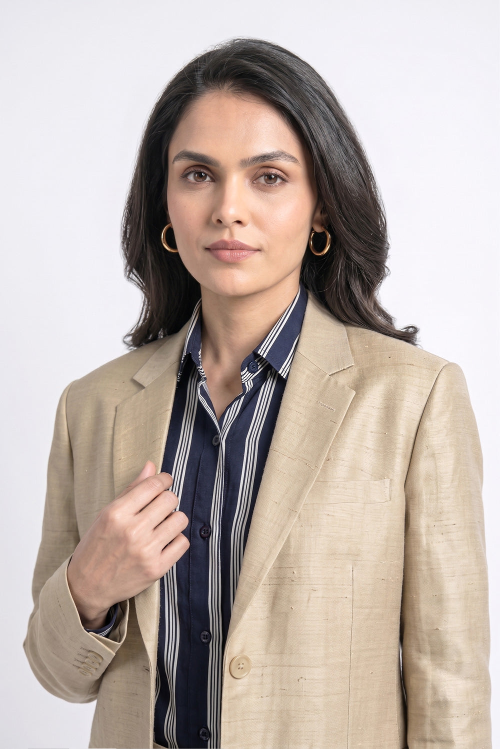 A woman wearing a beige textured silk blazer over a navy blue and white striped button-down shirt, with gold hoop earrings, photographed against a plain light gray studio background.