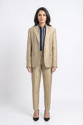 A woman stands against a light gray studio background wearing a tailored beige silkl blazer and matching trousers over a navy striped shirt, with black pointed heels.