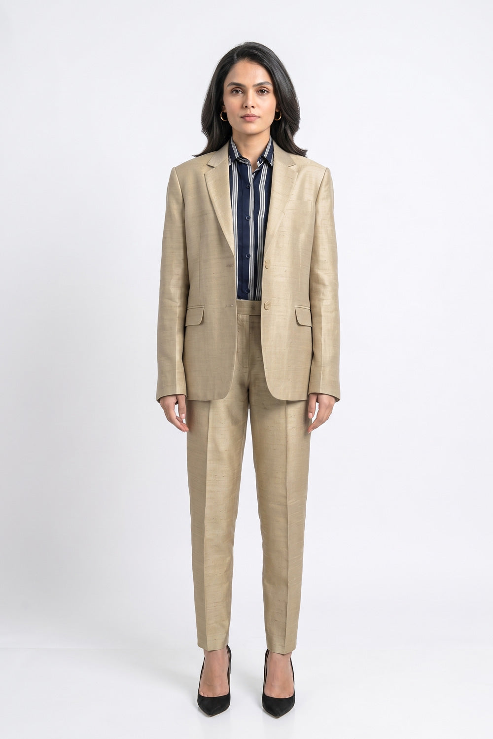 A woman stands against a light gray studio background wearing a tailored beige silkl blazer and matching trousers over a navy striped shirt, with black pointed heels.