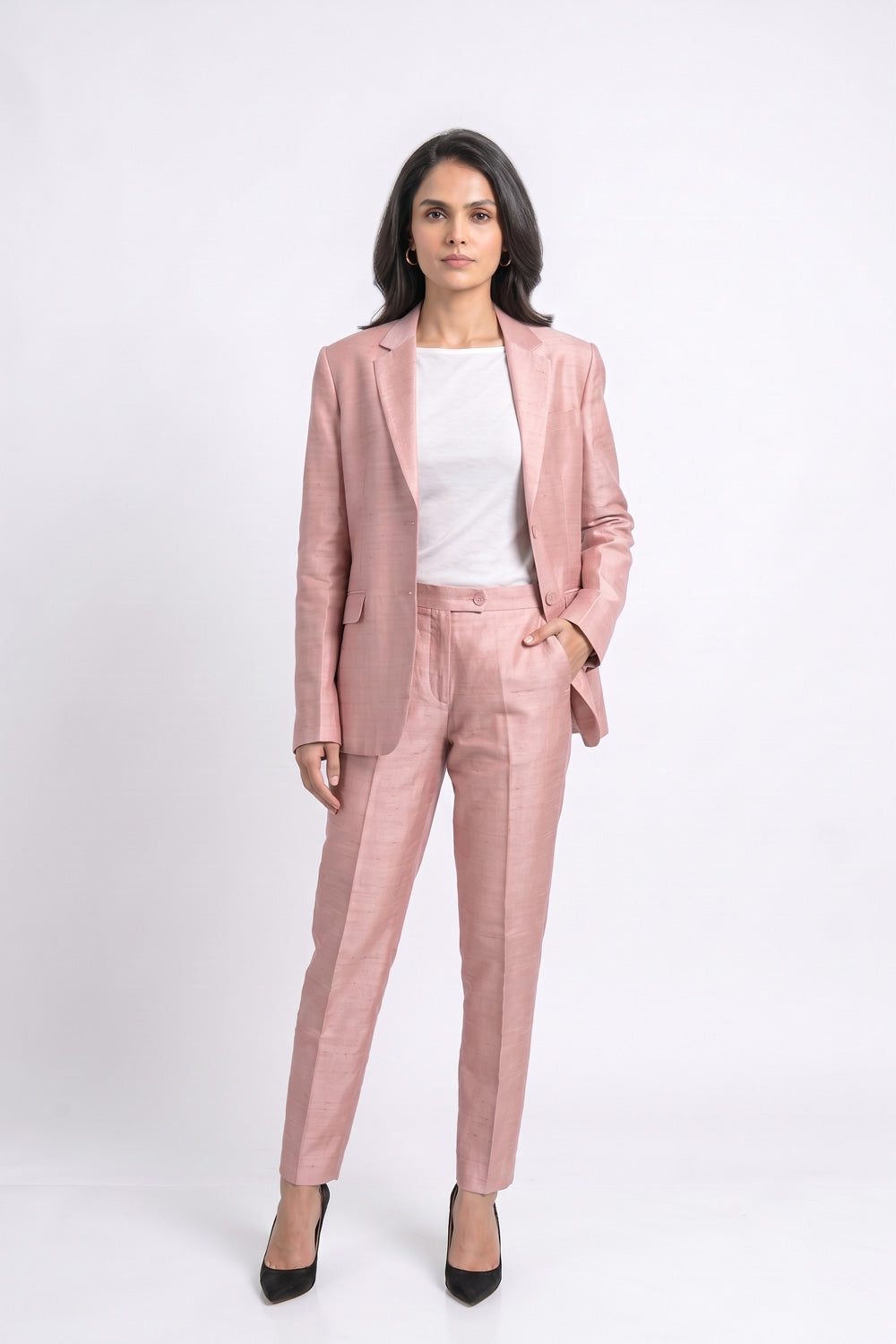 A woman modeling a dusty pink tailored suit set consisting of a silk blazer and matching trousers, styled with a white top and black heels against a light gray studio background.