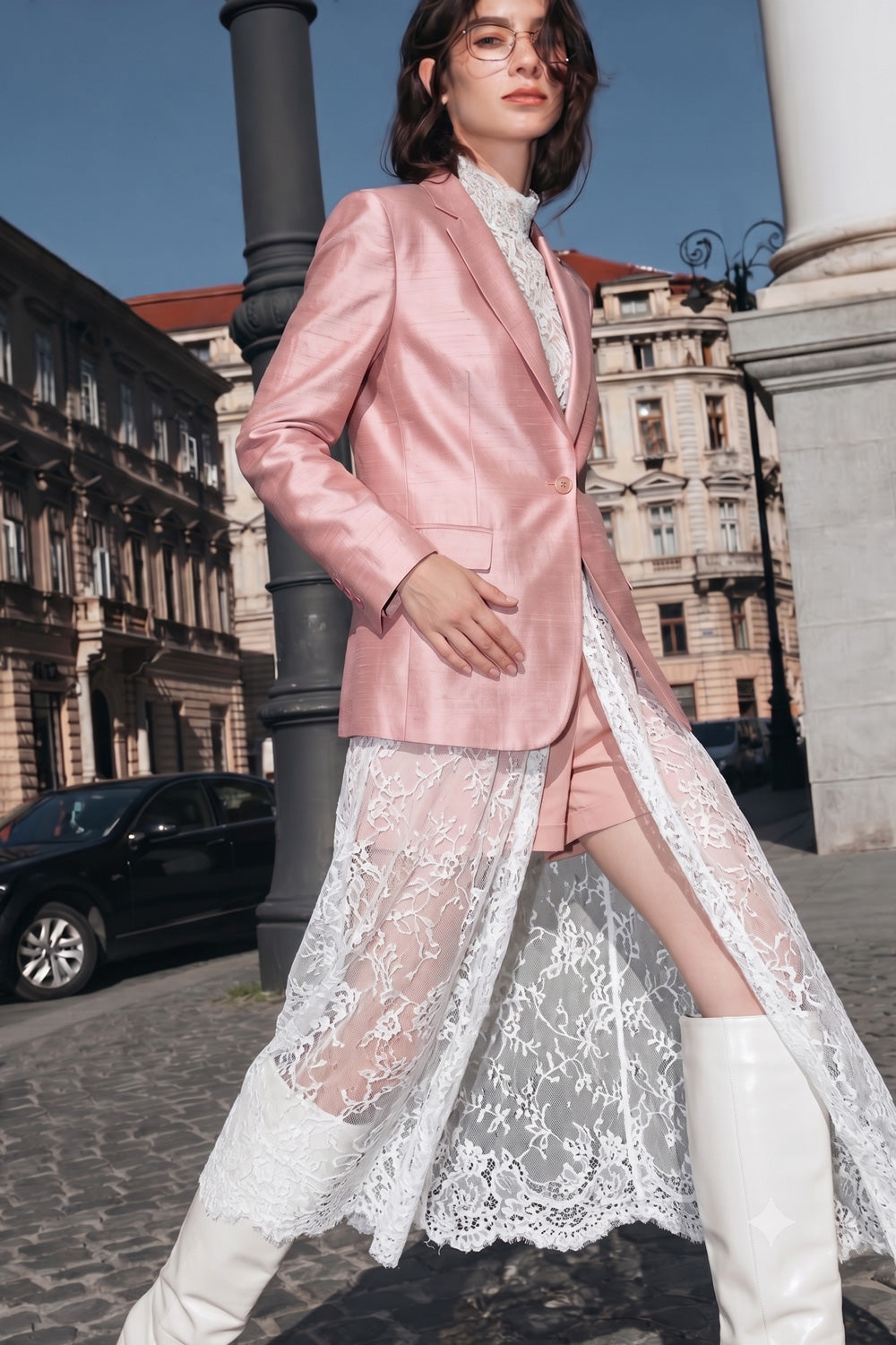 A model wearing a shiny light pink silk blazer, a white lace midi skirt, and white knee-high boots, standing in an outdoor city street with historic buildings in the background.