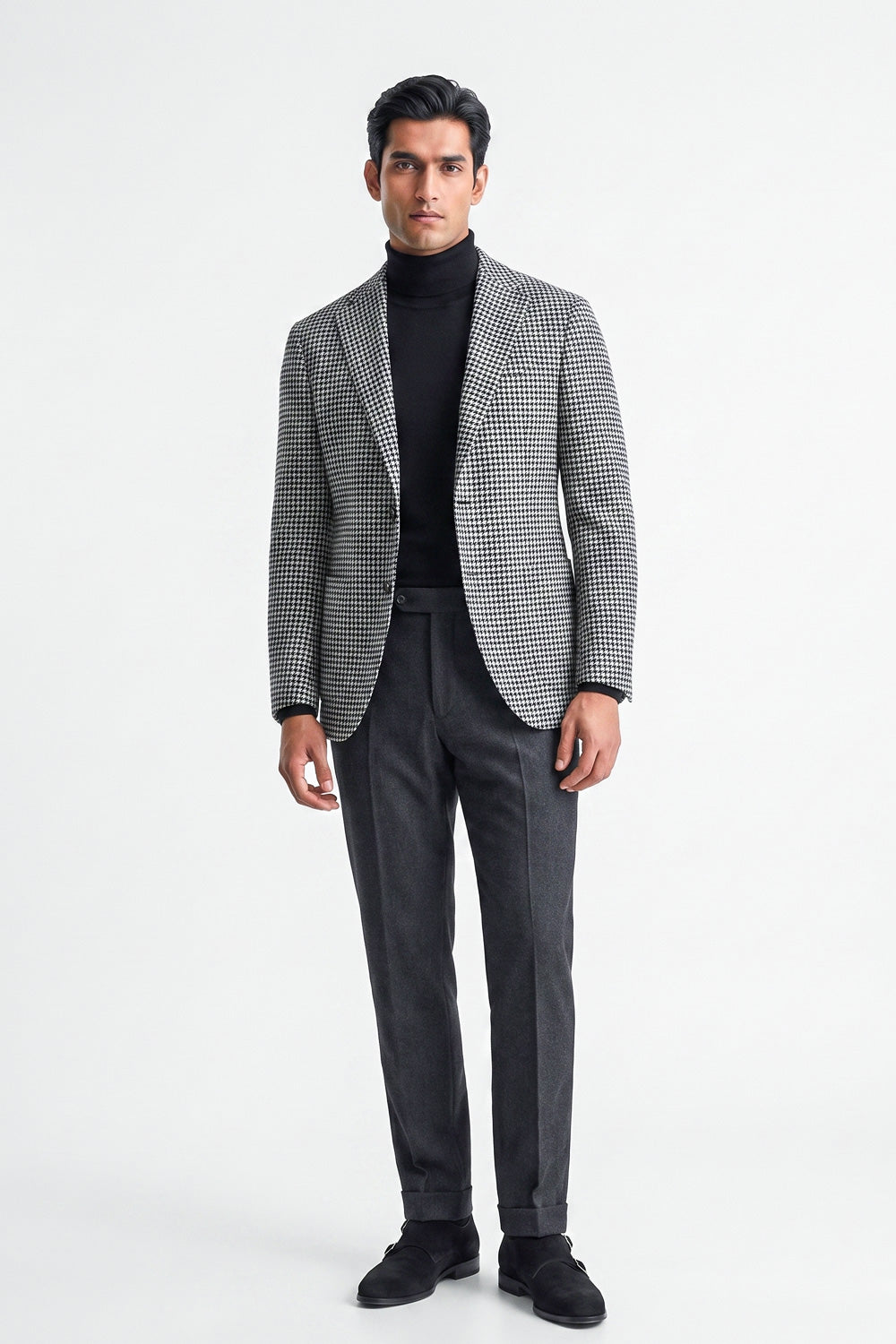 A man modeling a gray and black checks cotton blazer over a black turtleneck, paired with dark gray trousers and black loafers, against a plain white studio background.