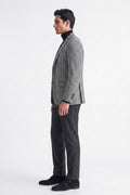 A man in profile wearing a black and white checks cotton blazer over a black turtleneck, paired with dark charcoal trousers and black loafers, standing against a plain white studio background.