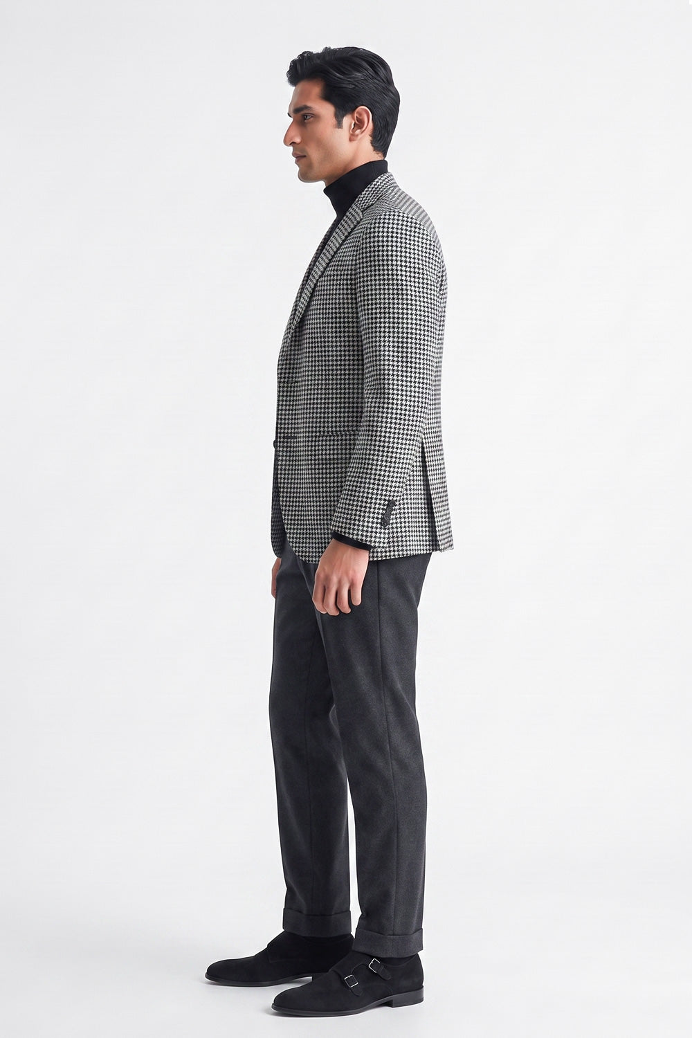A man in profile wearing a black and white checks cotton blazer over a black turtleneck, paired with dark charcoal trousers and black loafers, standing against a plain white studio background.
