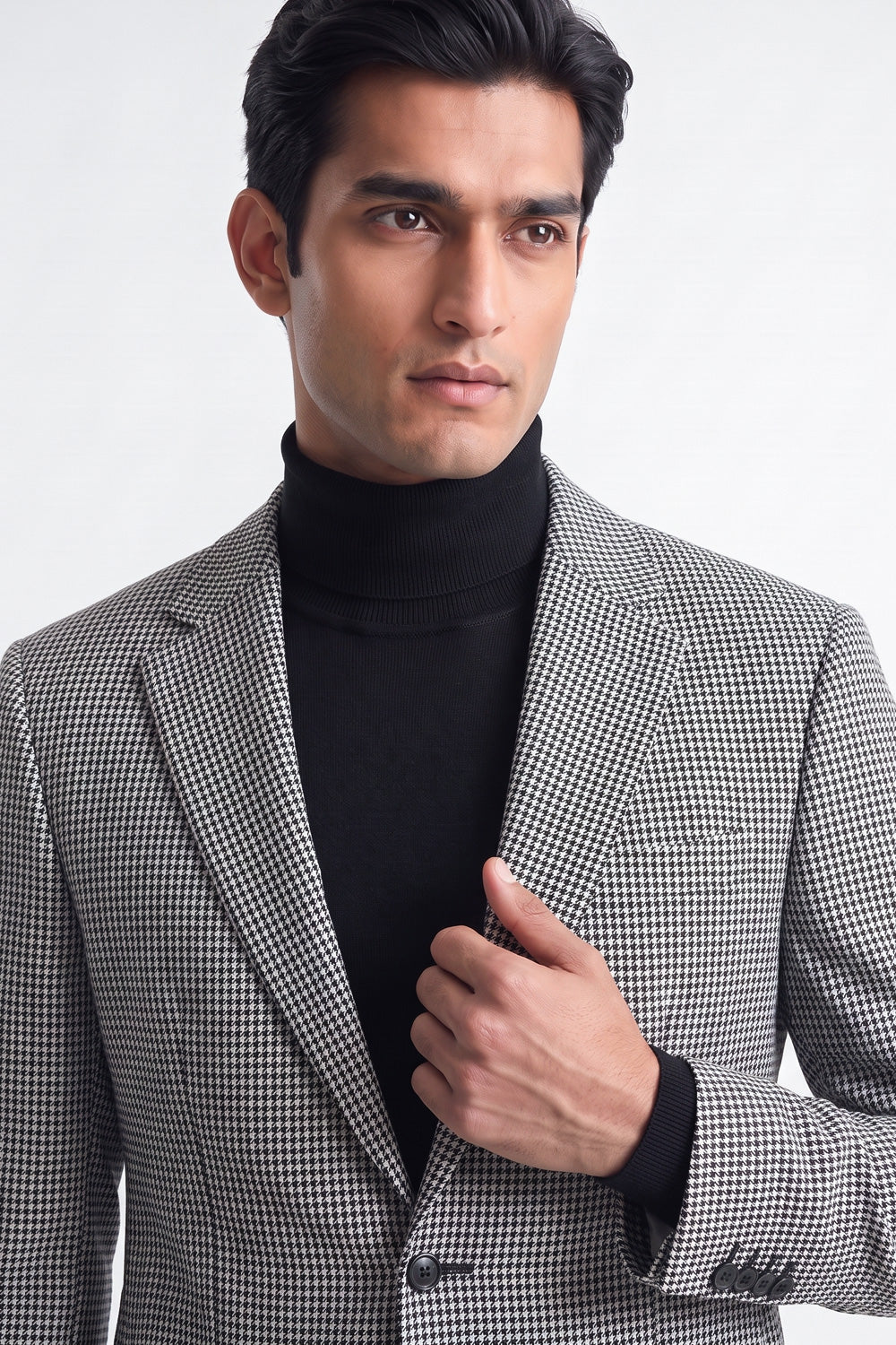 A man wearing a black and white checks cotton blazer over a black turtleneck sweater, photographed against a clean light gray studio background.