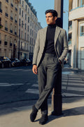 A man stands on a city street wearing a gray and black checks cotton blazer, a black turtleneck, gray trousers, and black shoes.
