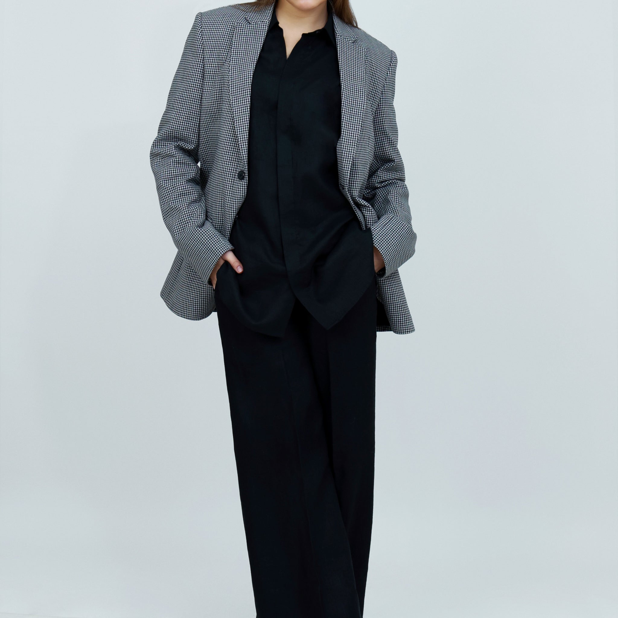 A person wearing a single-breasted, gray, checkered unisex cotton jacket with a notch lapel and functional sleeves, paired with a black outfit.