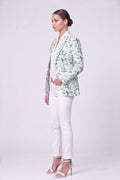 A woman stands in profile wearing a light-colored linen blazer with a blue and green floral pattern, white pants, and white high-heeled sandals against a plain light gray studio background.