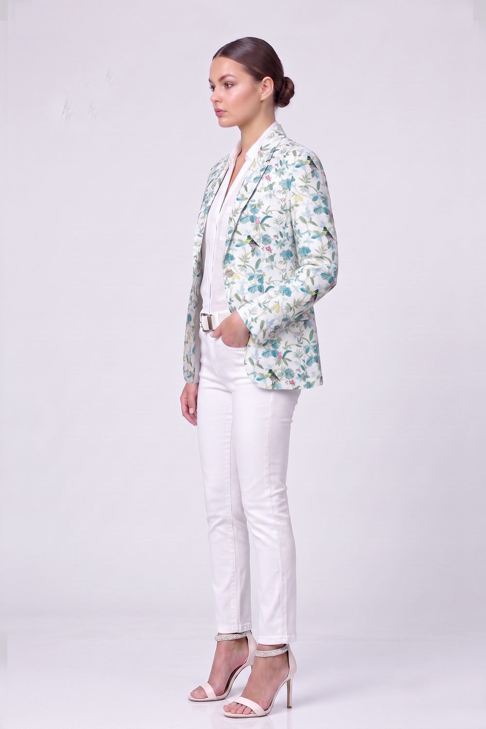 A woman stands in profile wearing a light-colored linen blazer with a blue and green floral pattern, white pants, and white high-heeled sandals against a plain light gray studio background.