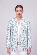 A woman modeling a white linen blazer with a colorful floral and bird pattern, worn over a white collared shirt and white trousers against a light gray studio background.