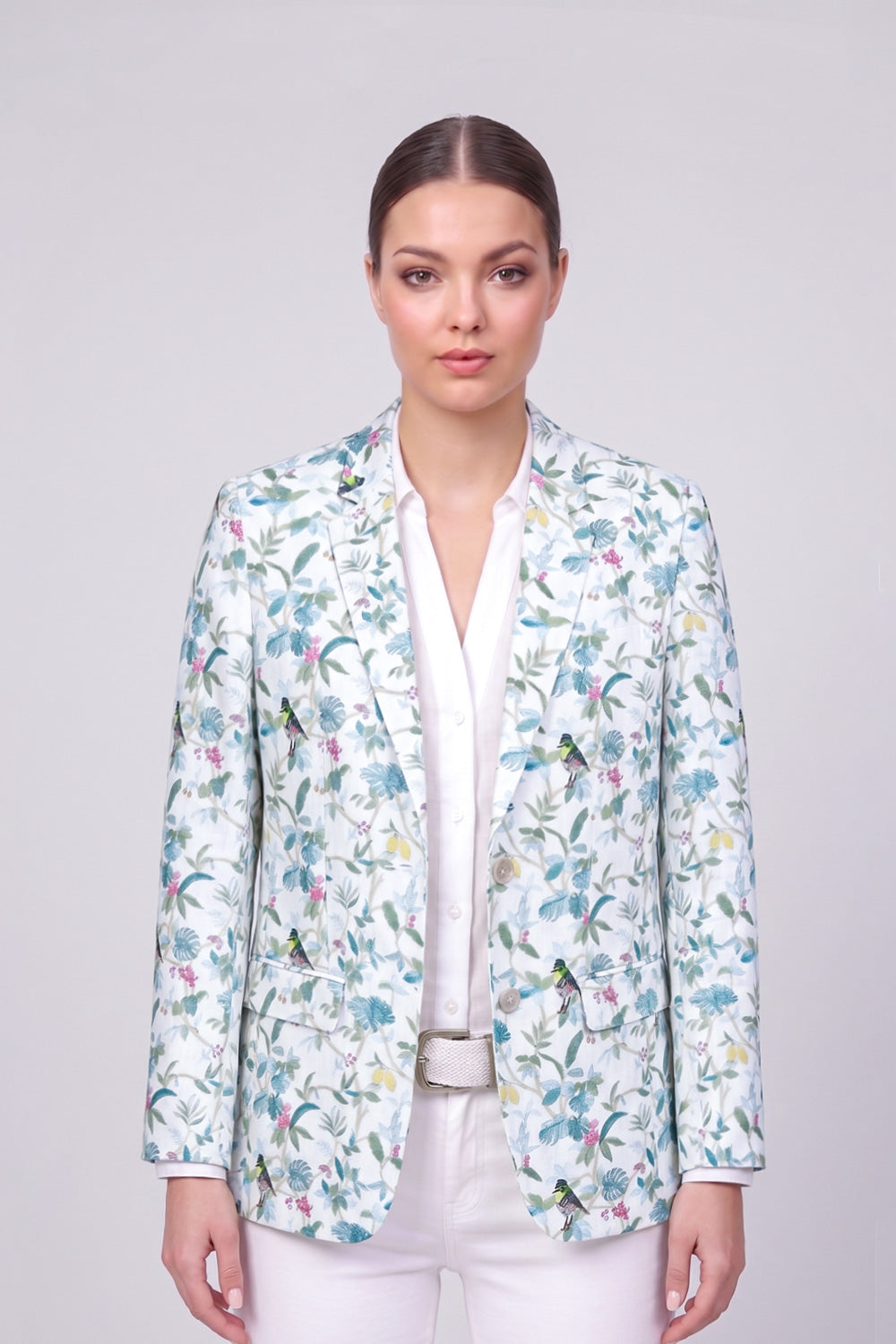 A woman modeling a white linen blazer with a colorful floral and bird pattern, worn over a white collared shirt and white trousers against a light gray studio background.