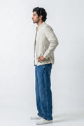 A man in profile wearing a light beige linen jacket over a white shirt, blue jeans, and white sneakers, standing against a plain white studio background.