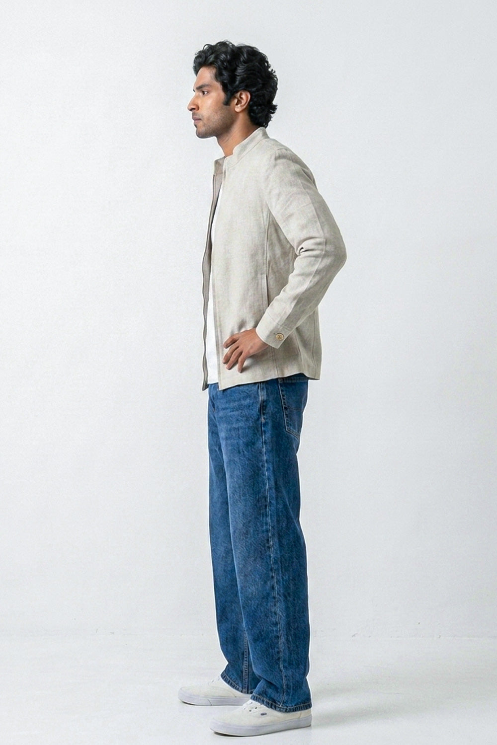 A man in profile wearing a light beige linen jacket over a white shirt, blue jeans, and white sneakers, standing against a plain white studio background.