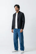 A man wearing a black zip-up linen jacket over a white t-shirt, blue jeans, and white sneakers, standing against a plain white studio background.