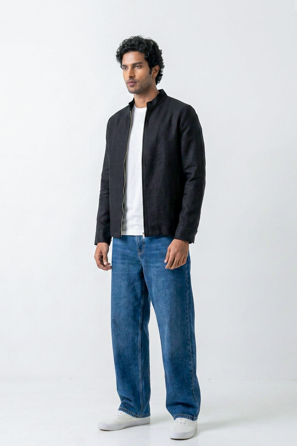 A man wearing a black zip-up linen jacket over a white t-shirt, blue jeans, and white sneakers, standing against a plain white studio background.
