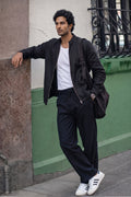 A man wearing a black bomber linen jacket, black trousers, a white t-shirt, and white sneakers with black stripes, leaning against a green and beige wall while carrying a black shoulder bag.