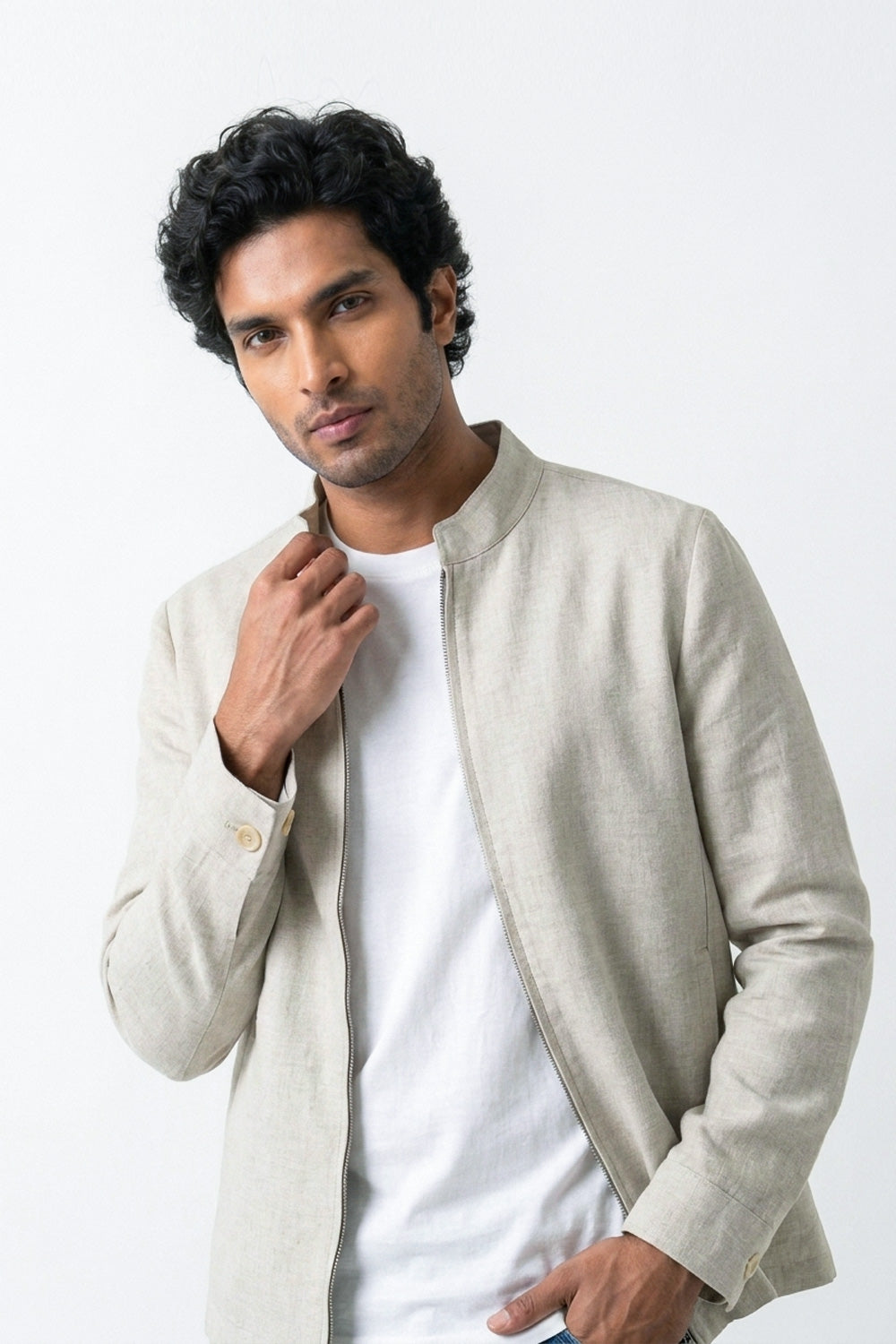 A man wearing a light beige linen-jacket over a white t-shirt, posing against a plain white studio background.