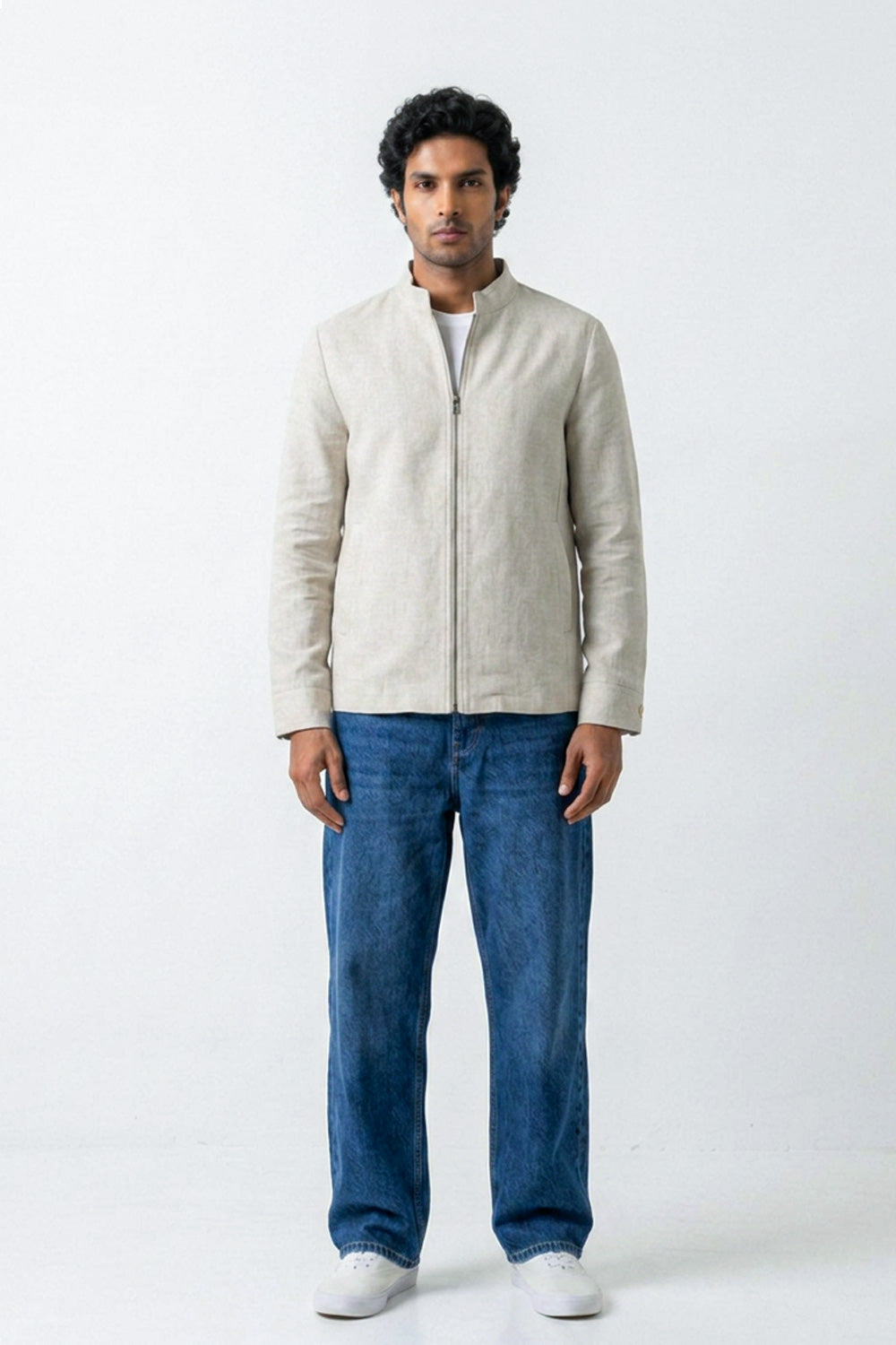 A man wearing a light beige linen jacket with a stand collar and front zipper, paired with blue jeans and white sneakers, standing against a white studio background.