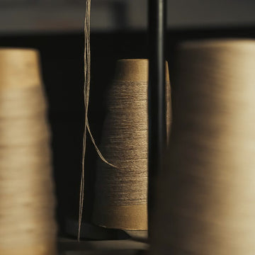 A close-up, artistic shot of a spool of beige linen thread wound on a cone, framed by blurred spools of matching thread in the foreground and background.