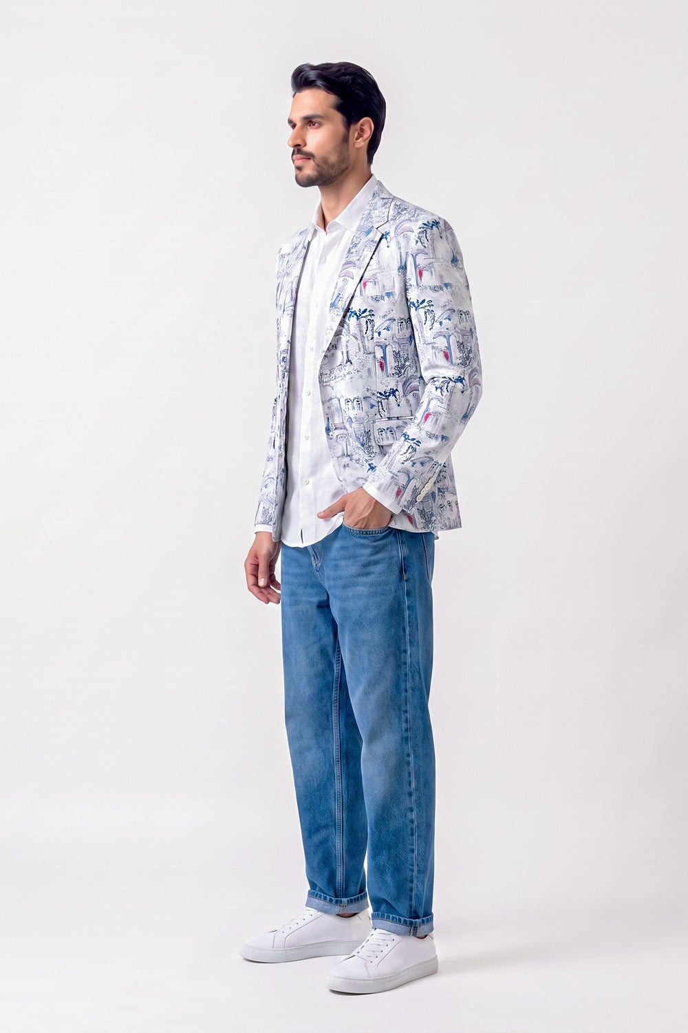 A man wearing a white linen blazer with a colorful graffiti-style print, a white t-shirt, blue jeans, and white sneakers, standing against a plain white studio background.