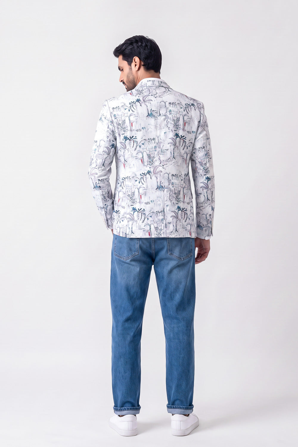 A man viewed from behind wearing a white long-sleeve linen blazer with a blue and gray tropical print, paired with blue jeans and white sneakers, against a plain light gray studio background.