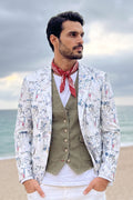 A man standing on a beach wearing a white patterned linen blazer over an olive green vest and white shirt, with a red neck scarf.