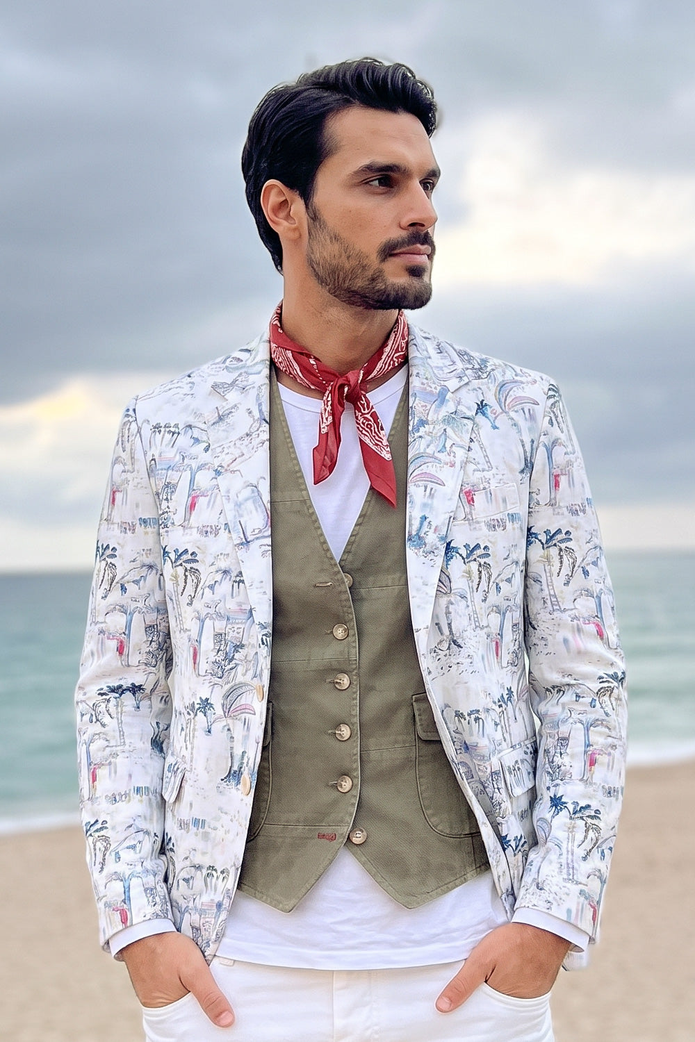 A man standing on a beach wearing a white patterned linen blazer over an olive green vest and white shirt, with a red neck scarf.