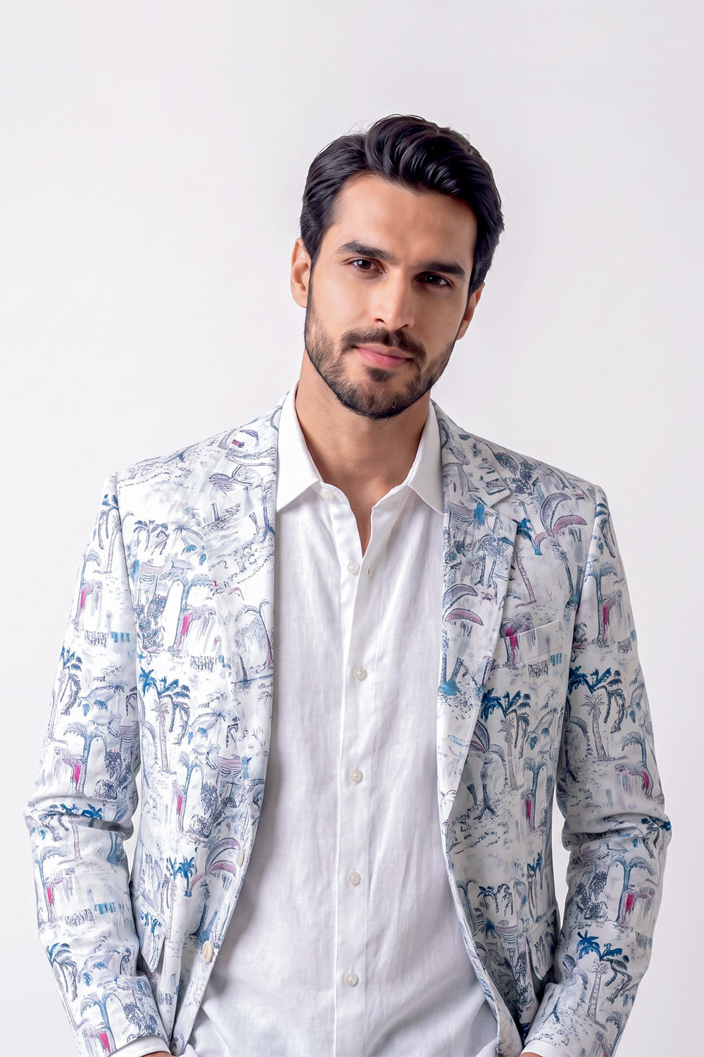 A man wearing a white button-up shirt and a patterned linen blazer with blue, pink, and gray tropical illustrations, posed against a light gray studio background.