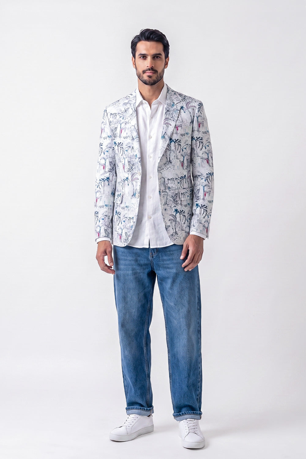 A man modeling a white linen blazer with a blue and gray toile-style print, worn over a white shirt and blue jeans, standing against a light gray studio background.