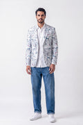 A man modeling a white linen blazer with a blue and gray toile-style print, worn over a white shirt and blue jeans, standing against a light gray studio background.