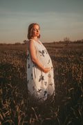 A pregnant woman stands in a field at sunset wearing a sleeveless white dress with black botanical print leaf patterns. The warm golden light illuminates the scene, and the sky is a soft blue-gray.